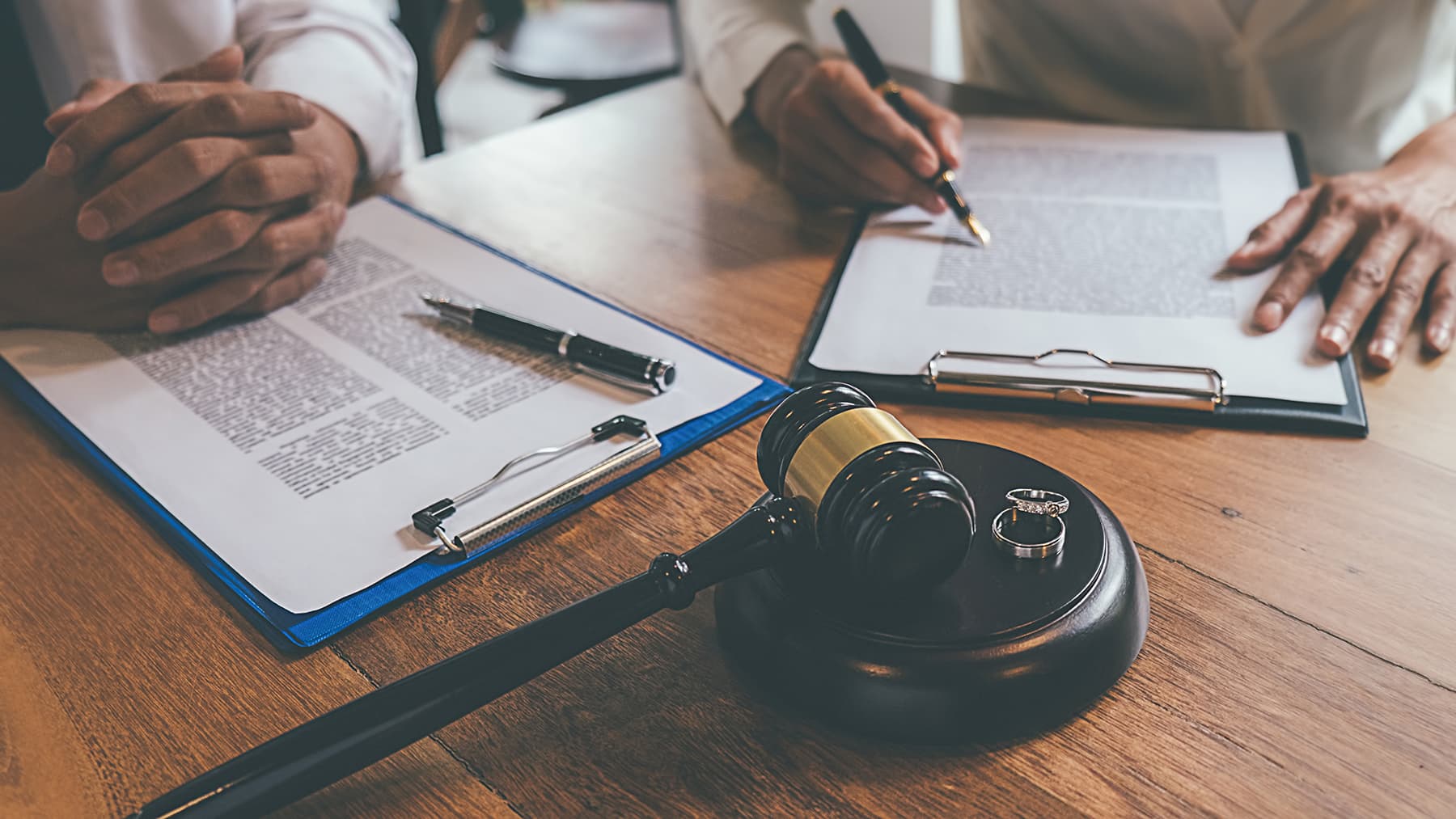 Two people sign legal documents at a wooden desk, with a judge's gavel and rings in view on the table.