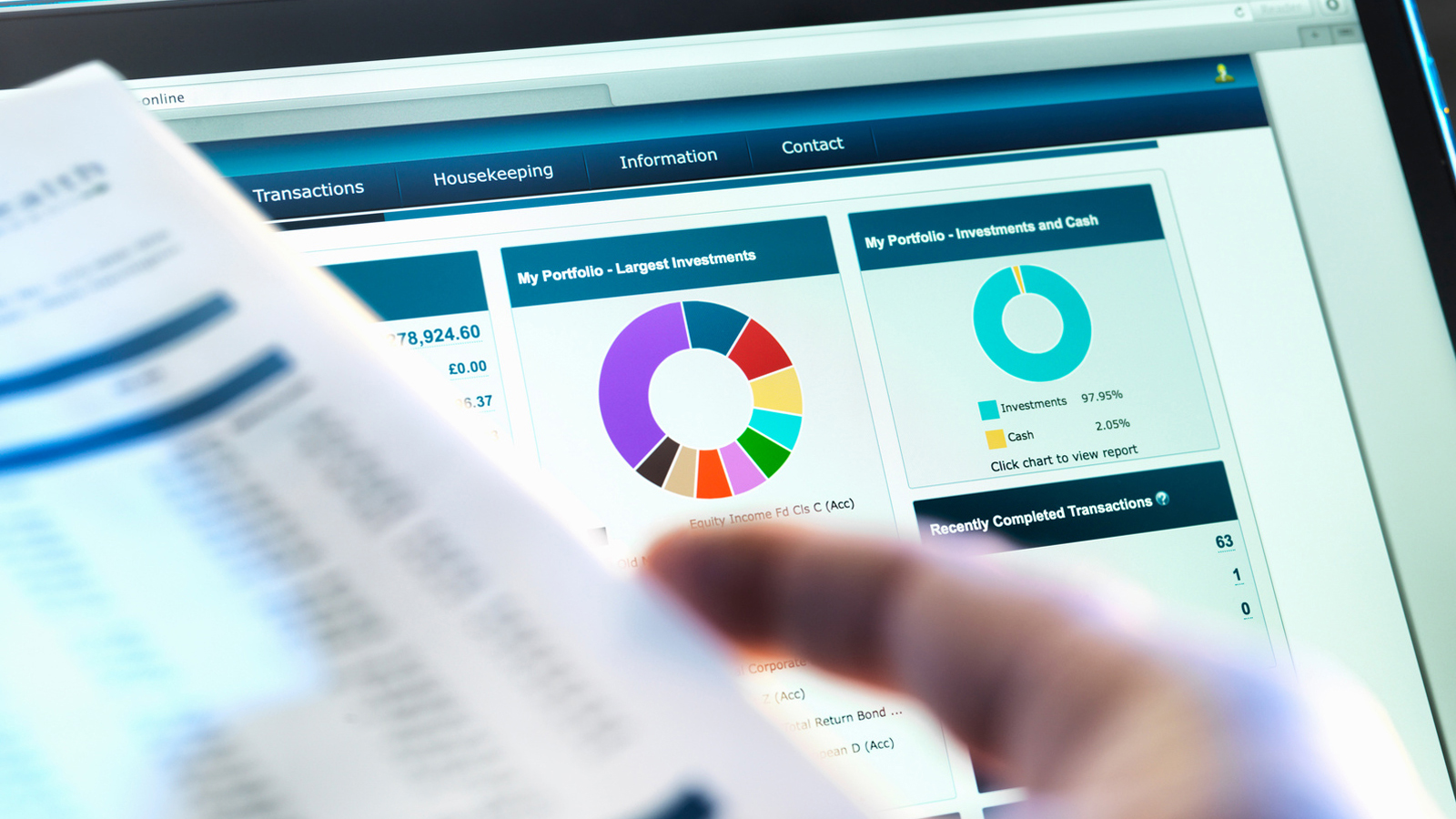 Person flipping a paper in front of a computer monitor displaying financial portfolio dashboards and charts