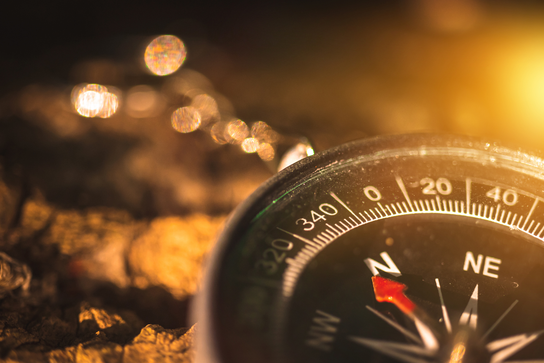Close-up of a compass resting on rough ground with warm, blurry bokeh lights in the background.