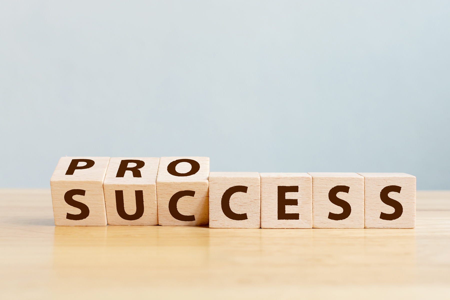 Wooden letter blocks spell out 'PROCESS SUCCESS' on a light desk backdrop, conveying achievement goals.