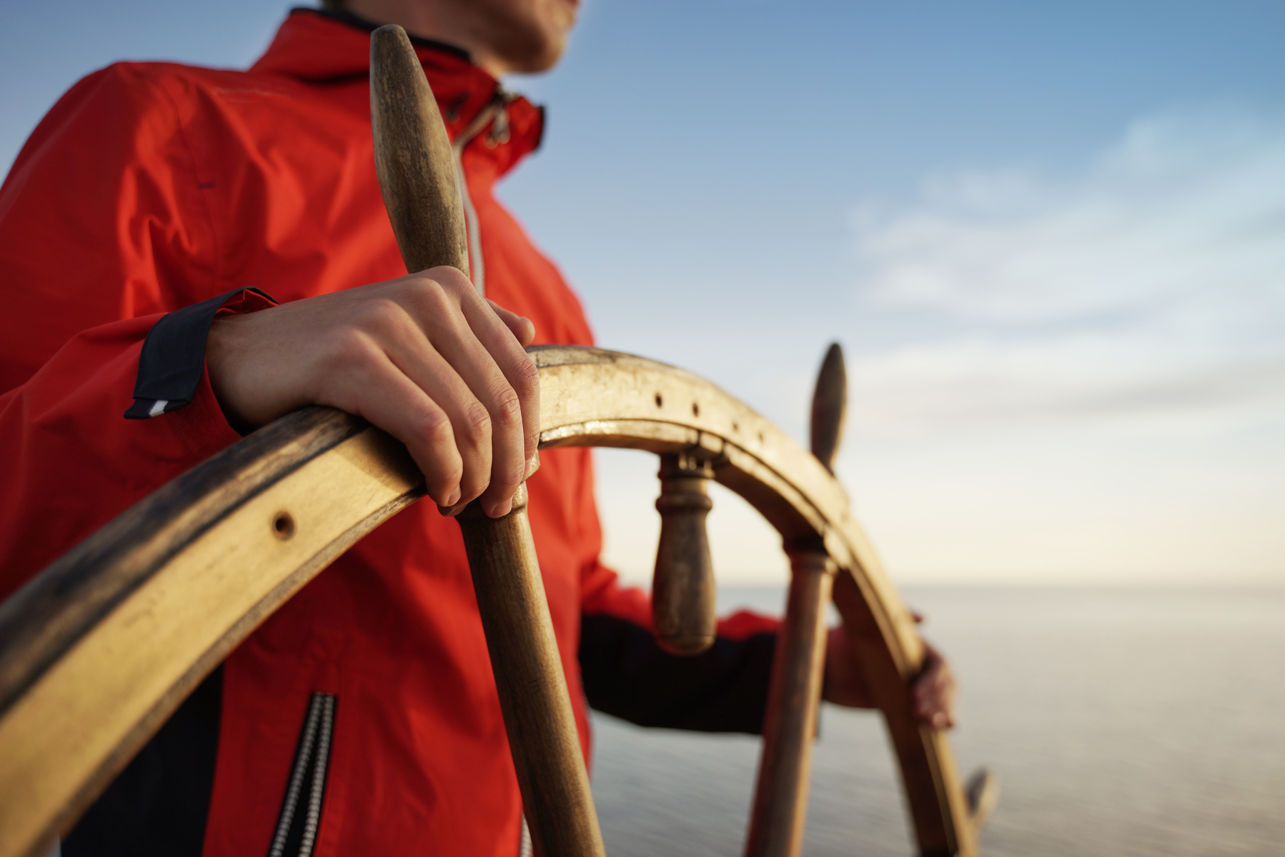Close-up of a person in a red jacket gripping a wooden ship's wheel at sea.