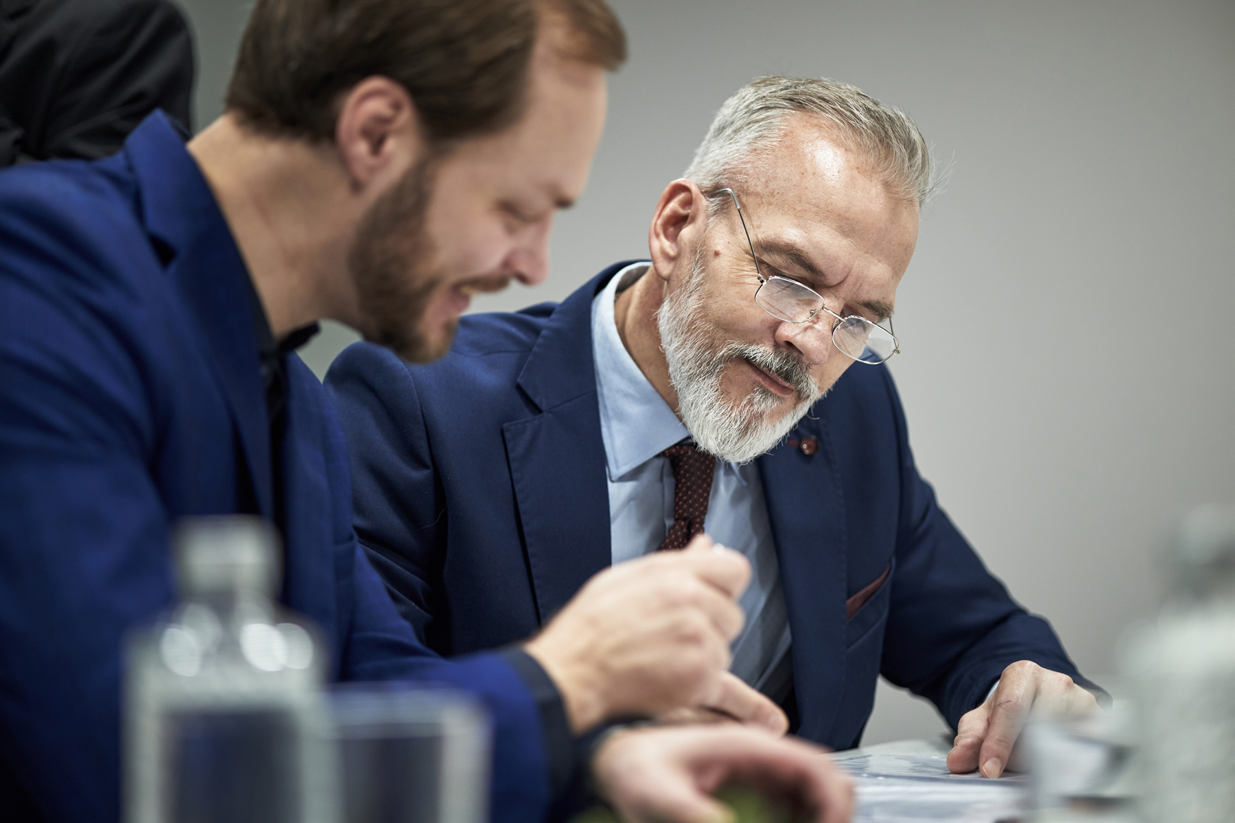 Two businessmen in blue suits sit at a table reviewing documents together.