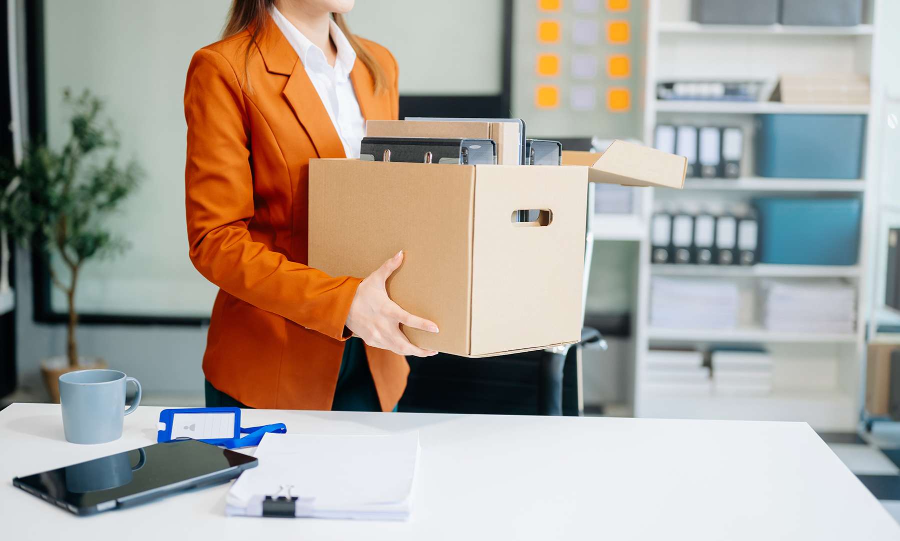 Woman in an orange blazer carries a cardboard box filled with files in a bright office.