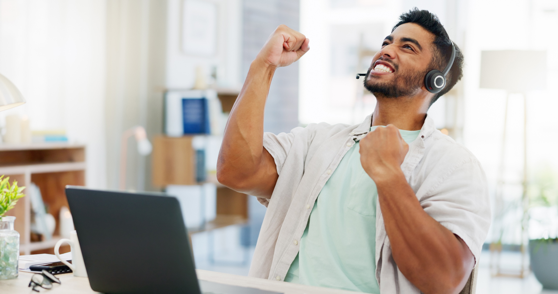 Man wearing a headset at a laptop, fist raised in celebration in a bright office setting