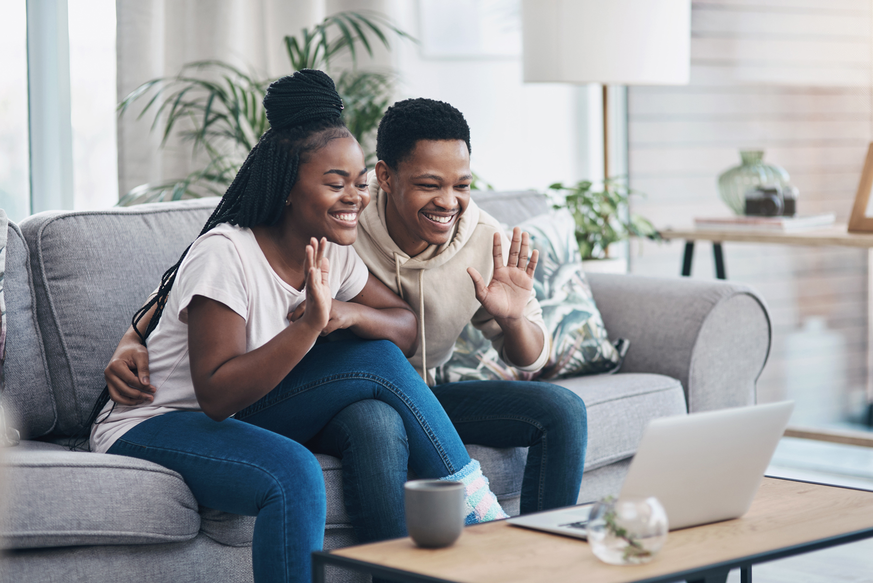 Two friends on a gray sofa smiling and waving at a laptop screen during a video call at home.